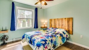 Ein Schlafzimmer mit einem Bett, einem Deckenventilator, einem Fenster mit Vorhängen, einem Nachttisch mit Lampe und einem an der Wand befestigten Holz-Kopfteil.