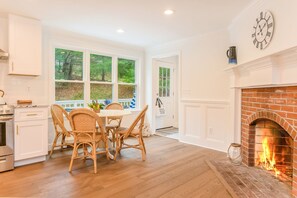 A kitchen with a dining area, a brick fireplace, and a large window overlooking greenery.