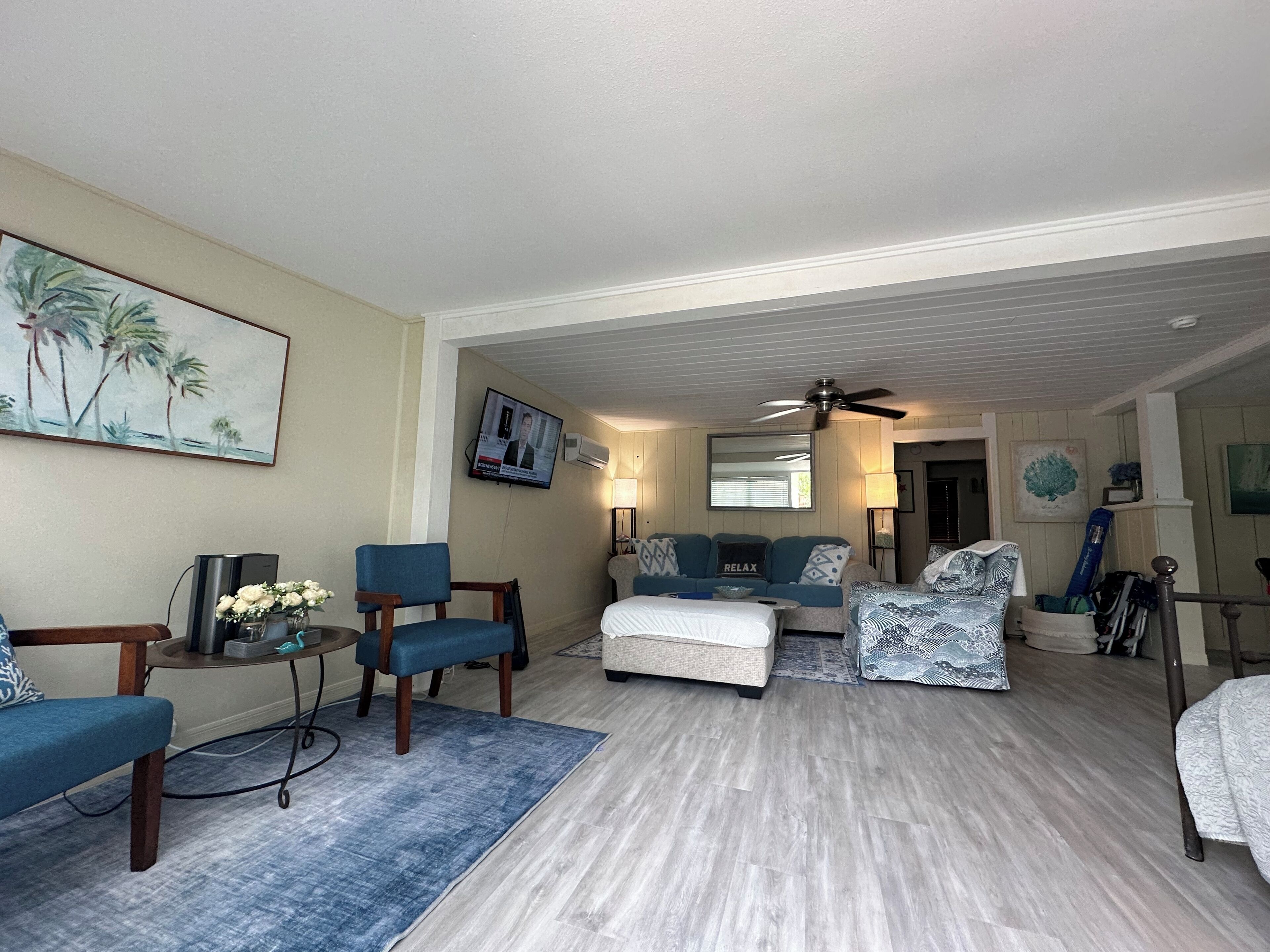 A spacious living room with a ceiling fan, a television, and a large painting of palm trees.