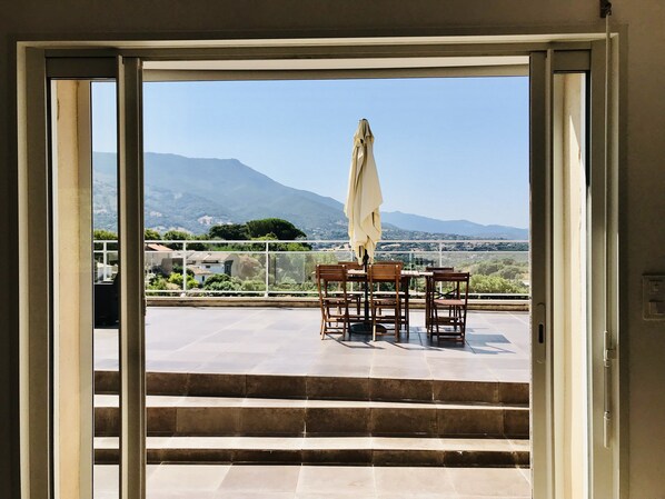 A view from a balcony with a table and chairs, a white umbrella, and a mountainous landscape in the background.