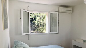 A bedroom with a bed, a window with open shutters, an air conditioning unit, and a bedside table. - Beautiful apartment on 1st floor of a quiet villa in the countryside, near Ajaccio (Sarrola-Carcopino)