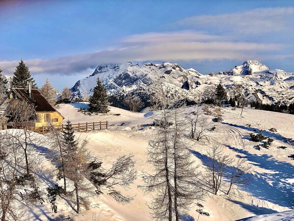 Paisaje nevado con una cabaña de madera y montañas al fondo.