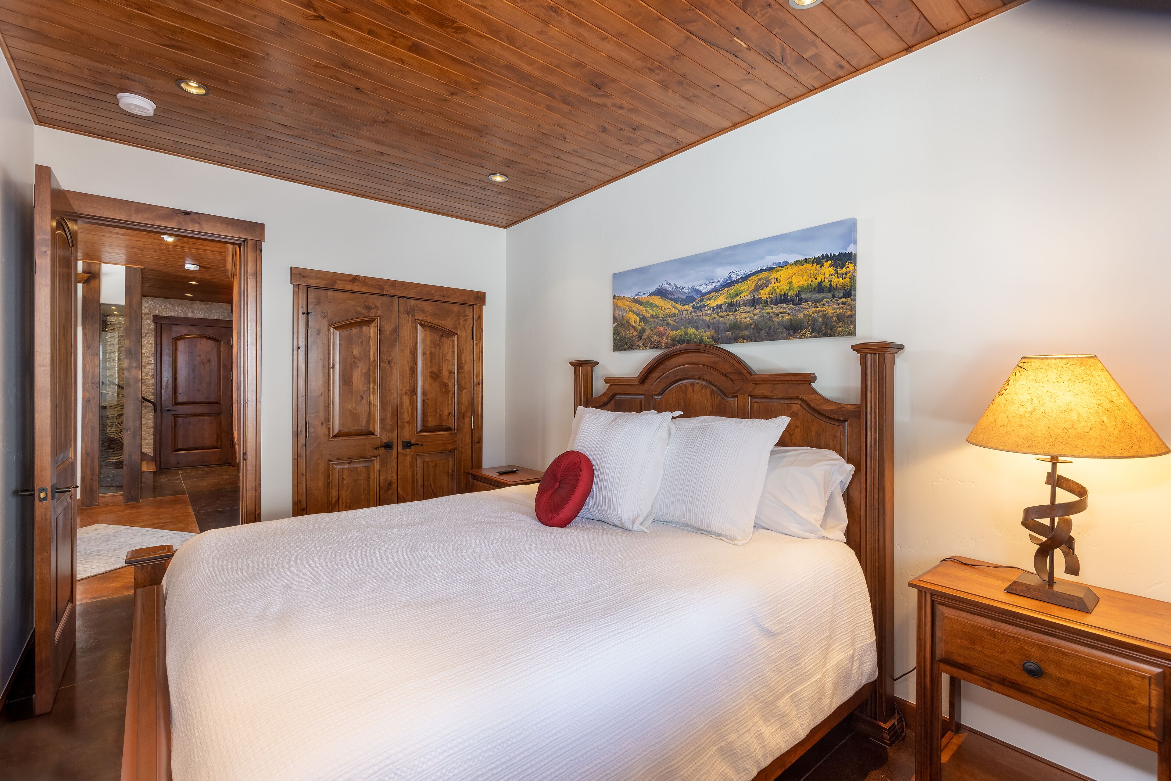 A bedroom with a wooden ceiling, a bed with white bedding, a red pillow, a wooden nightstand with a lamp, and a painting of a mountain landscape on the wall.