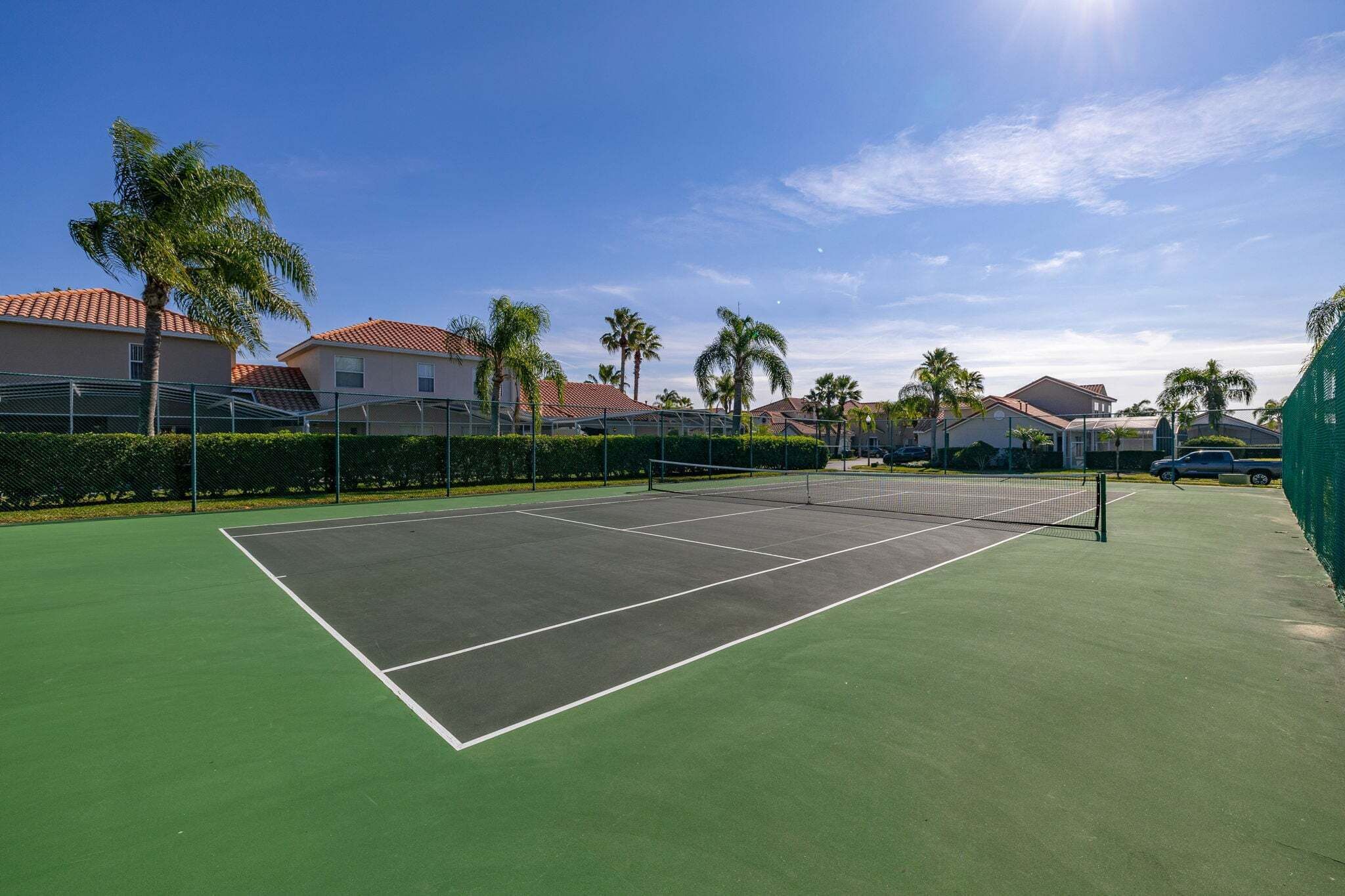 A tennis court with a net, surrounded by a fence, palm trees, and houses in the background.