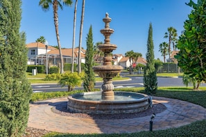 A fountain with multiple tiers, surrounded by a circular brick pathway, palm trees, and residential buildings in the background.