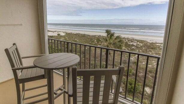 A balcony with a table and two chairs, overlooking a beach with dunes and a palm tree. - Oceanfront, Pet-Friendly Condo with Private Balcony! (St. Augustine)