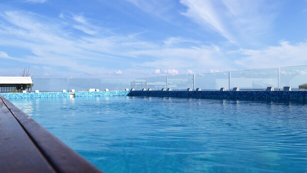 A clear blue pool with a wooden deck edge, surrounded by glass railings and a clear sky. - Pura Playa 501 by Riviera Rentals (Playa del Carmen)