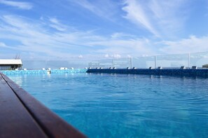 Une piscine d’un bleu éclatant, bordée d’une terrasse en bois, entourée de rampes de verre et surplombée par un ciel dégagé.