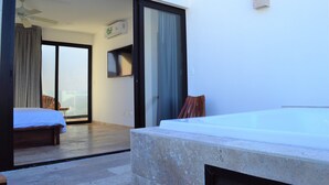 A modern hotel room with a large sliding glass door leading to a balcony, a wooden bed, a small table, and a hot tub. - Pura Playa 501 by Riviera Rentals (Playa del Carmen)