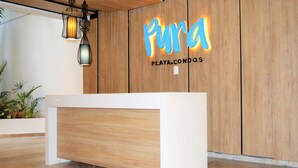 A reception desk with a wooden finish, a modern light fixture, and a wall with the name 'Pura' and 'PLAYA.CONDO'S' illuminated. - Pura Playa 501 by Riviera Rentals (Playa del Carmen)