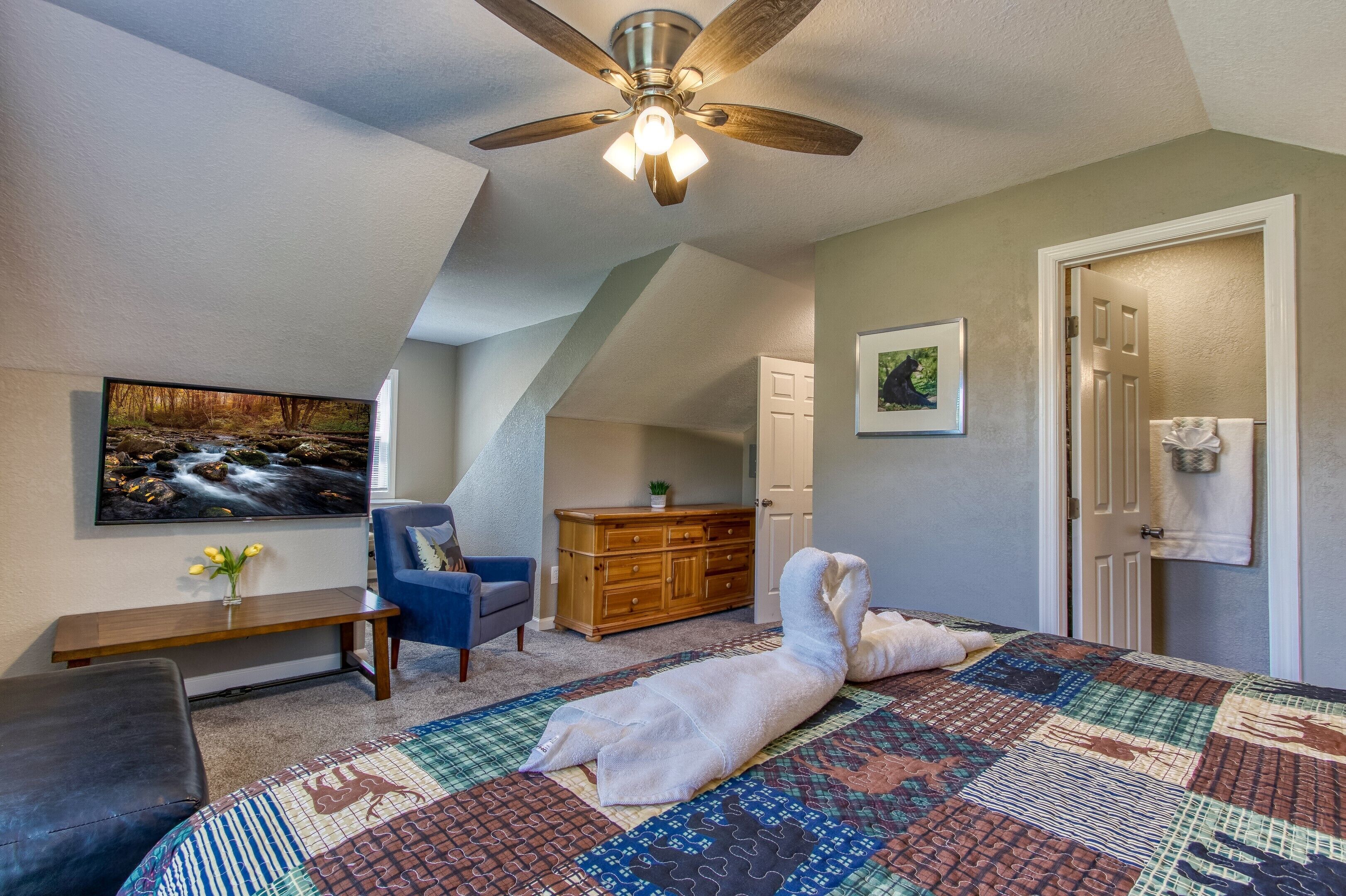 Une chambre avec un grand lit, un ventilateur de plafond, une télévision, une commode en bois, une chaise bleue et un tableau accroché au mur.