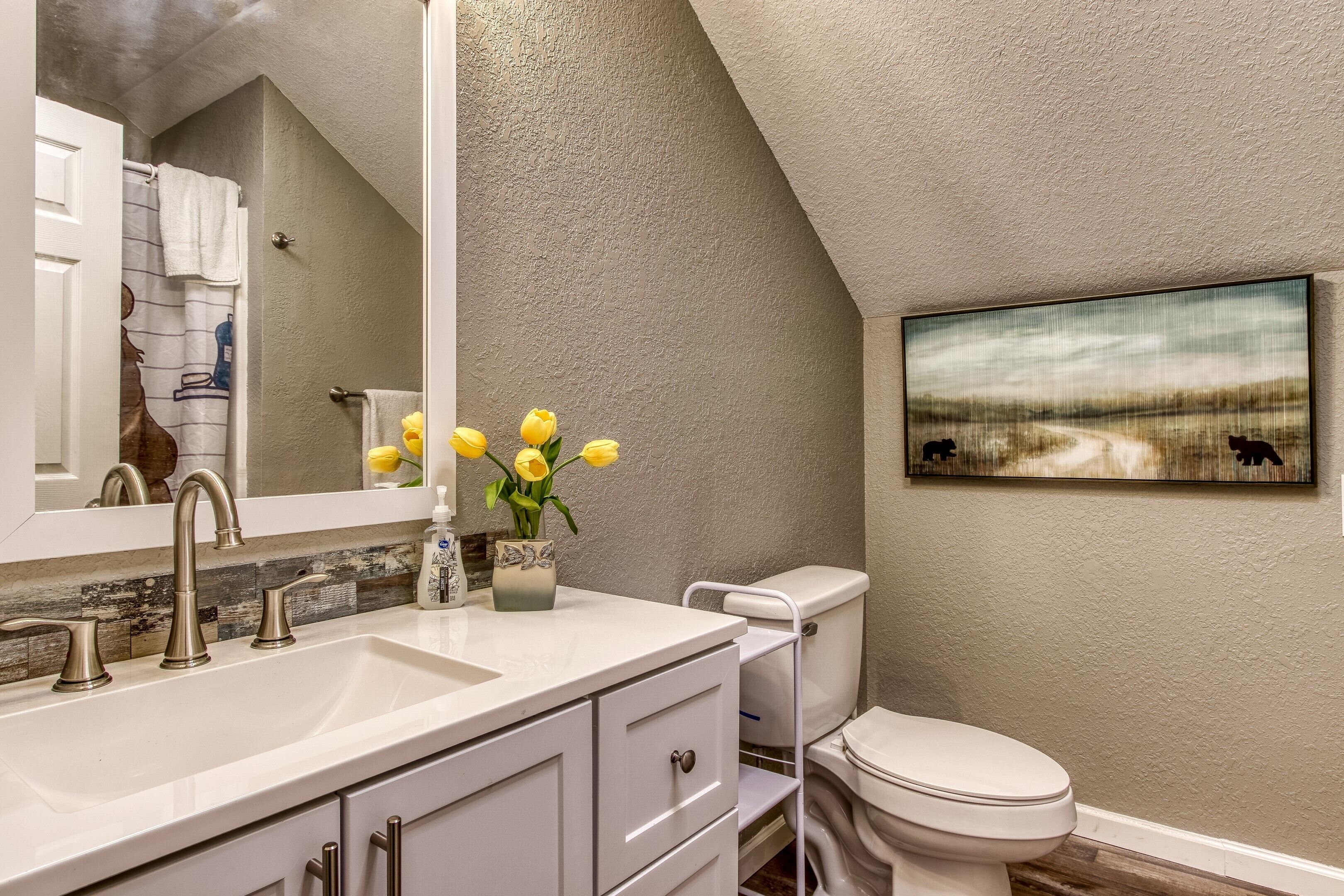 Une salle de bain avec un lavabo blanc, deux robinets, des toilettes et un grand miroir. Un vase rempli de fleurs jaunes est posé sur le comptoir du lavabo.