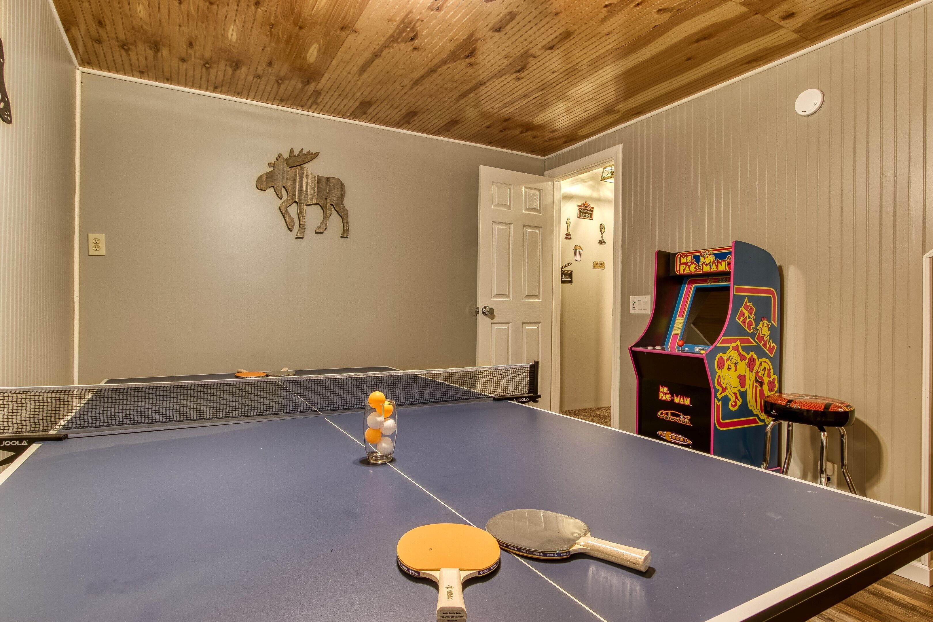 Une table de ping-pong avec deux raquettes et un porte-balles, un plafond en bois et une borne d’arcade rétro.