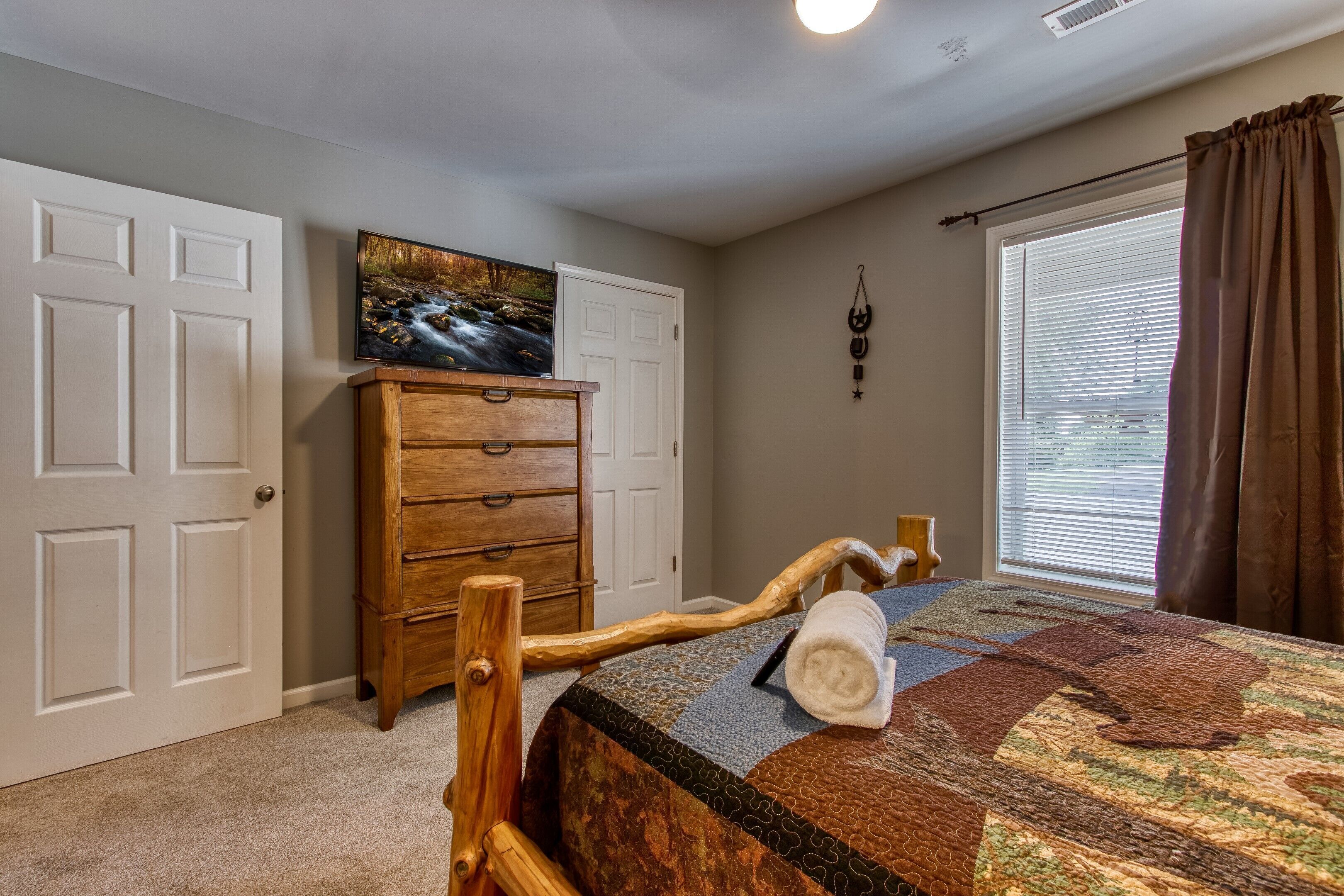A bedroom with a wooden bed frame, a quilted bedspread, a wooden dresser, a painting, and a window with blinds.