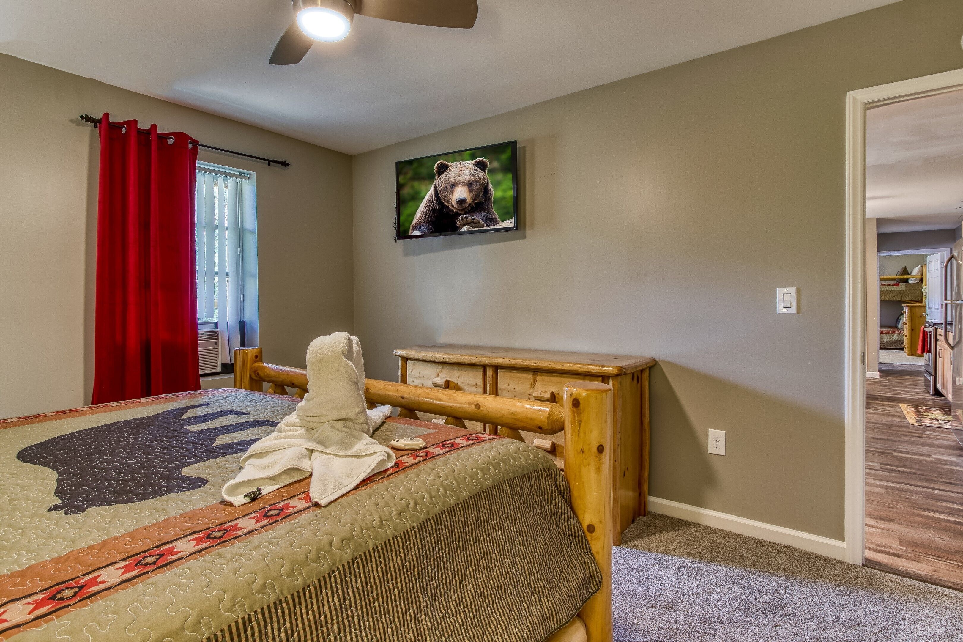 A bedroom with a bed, a wooden nightstand, a wall-mounted TV, and a window with red curtains.