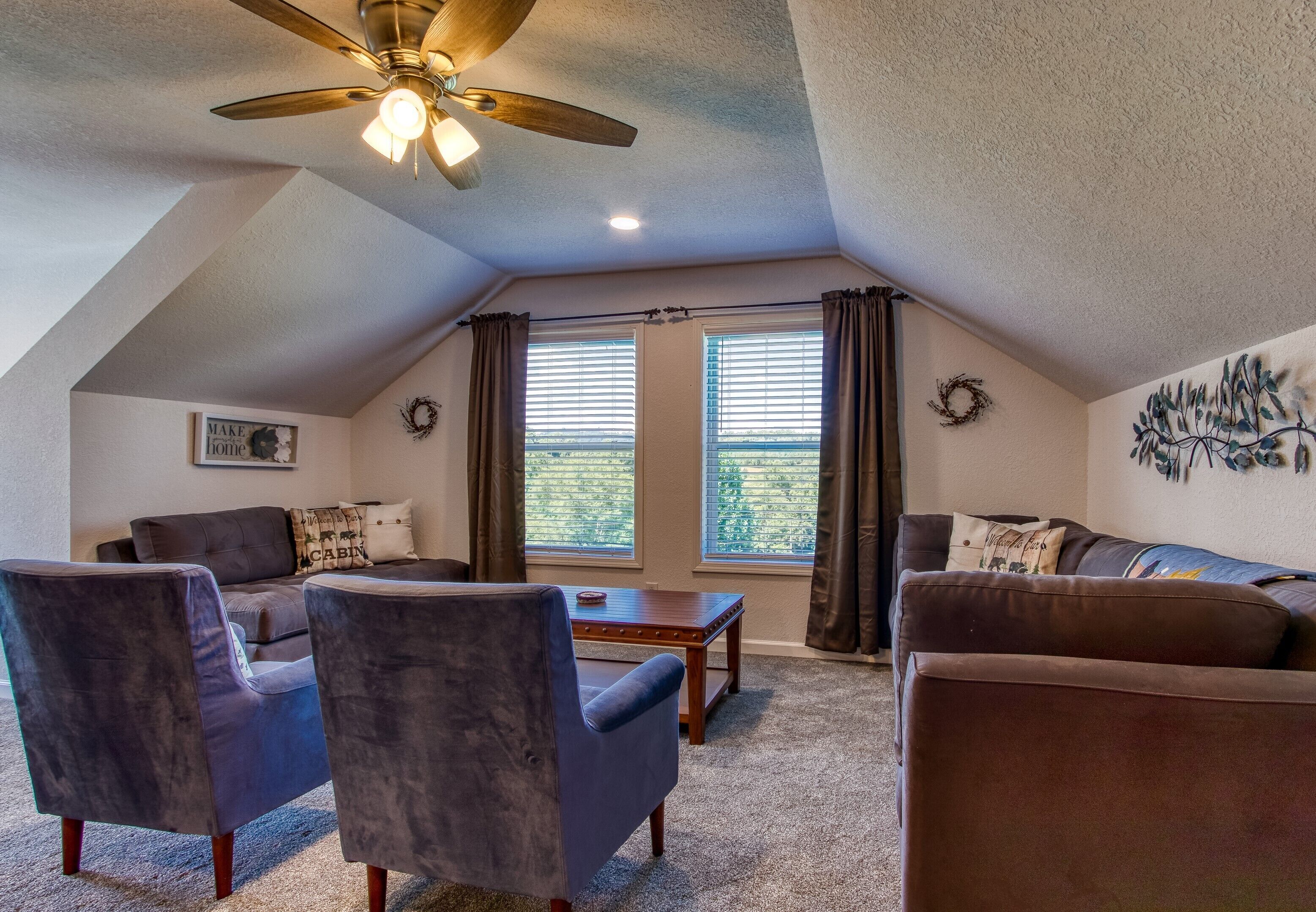 A cozy loft-style living room with a ceiling fan, two windows with curtains, a fireplace, and a wall-mounted artwork.