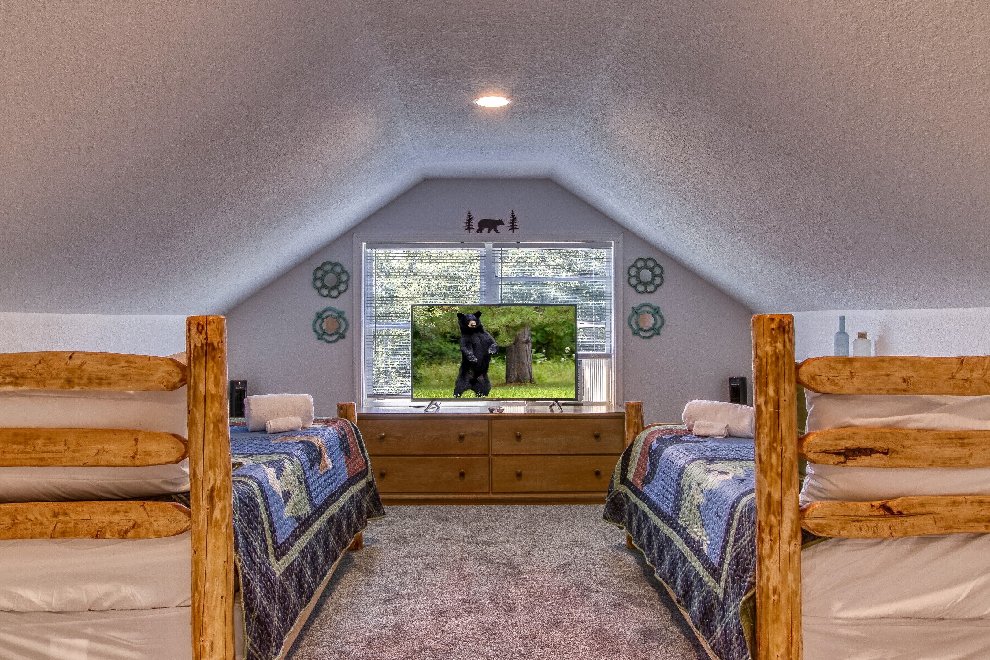 Une chambre avec deux lits superposés en bois, une grande fenêtre donnant sur les arbres, une télévision et une décoration représentant un ours sur le mur.