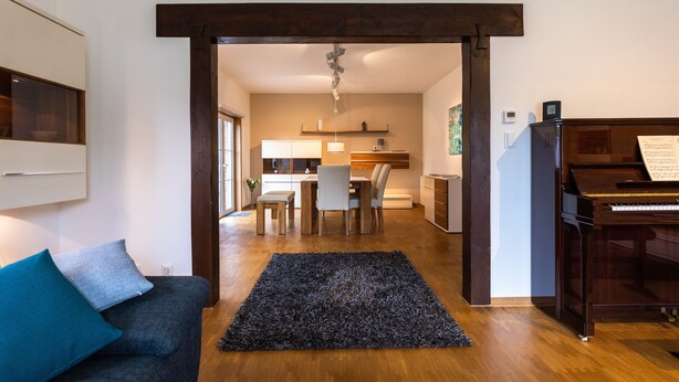 A living room with a dark grey sofa, a black shag rug, a wooden dining table with chairs, a piano, and a kitchen area in the background.