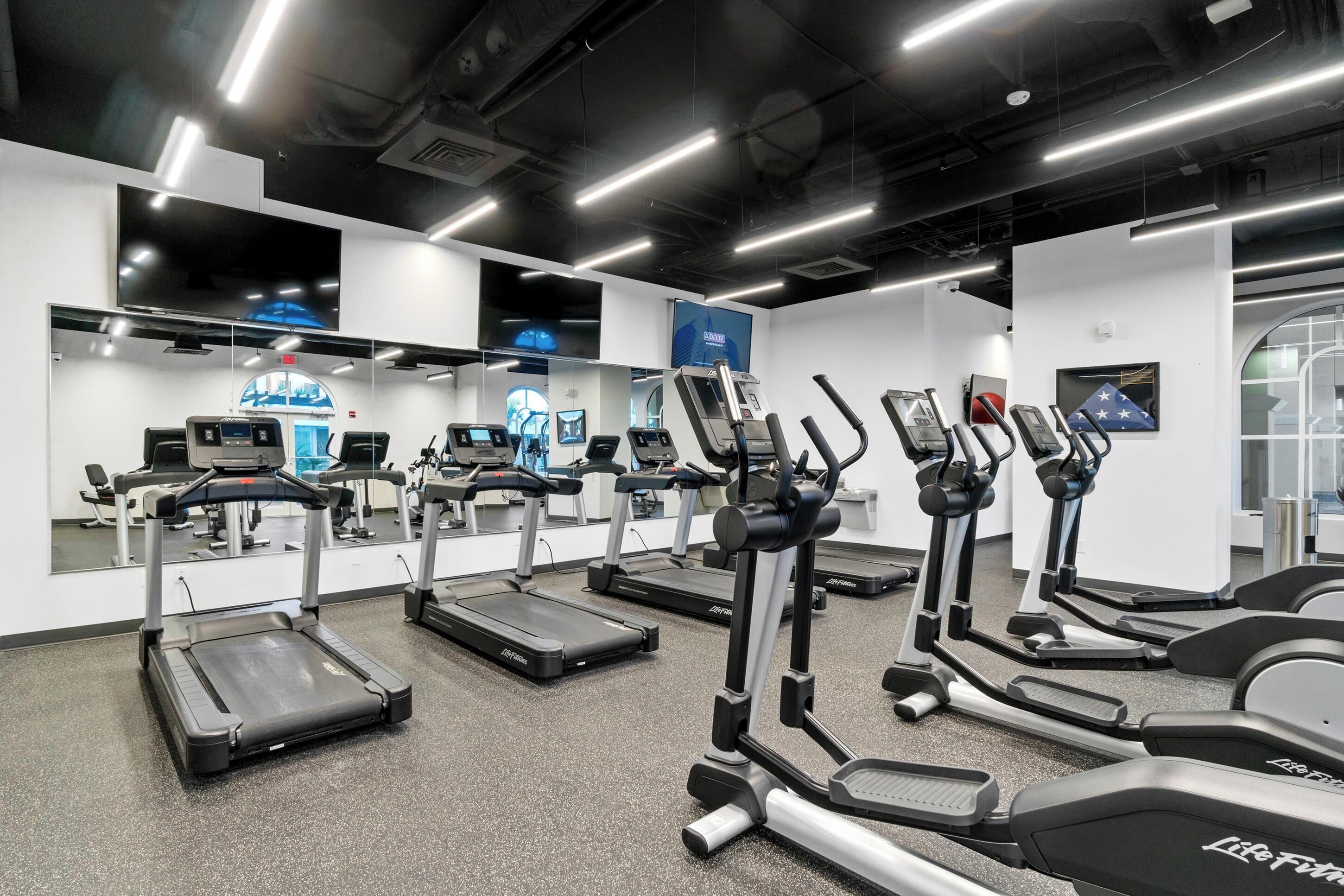 A gym with treadmills, ellipticals, and stationary bikes, equipped with mirrors and a large TV screen.