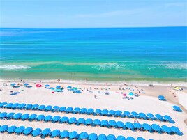 Ein Strand mit Reihen blauer Sonnenschirme und Liegestühlen, ein sandiger Uferstreifen und das Meer.