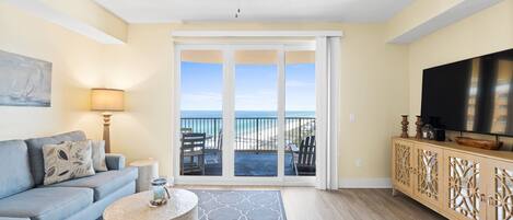 Ein Wohnzimmer mit einer Couch, einem runden Couchtisch, einem Fernseher und einem Balkon mit Blick auf den Strand.