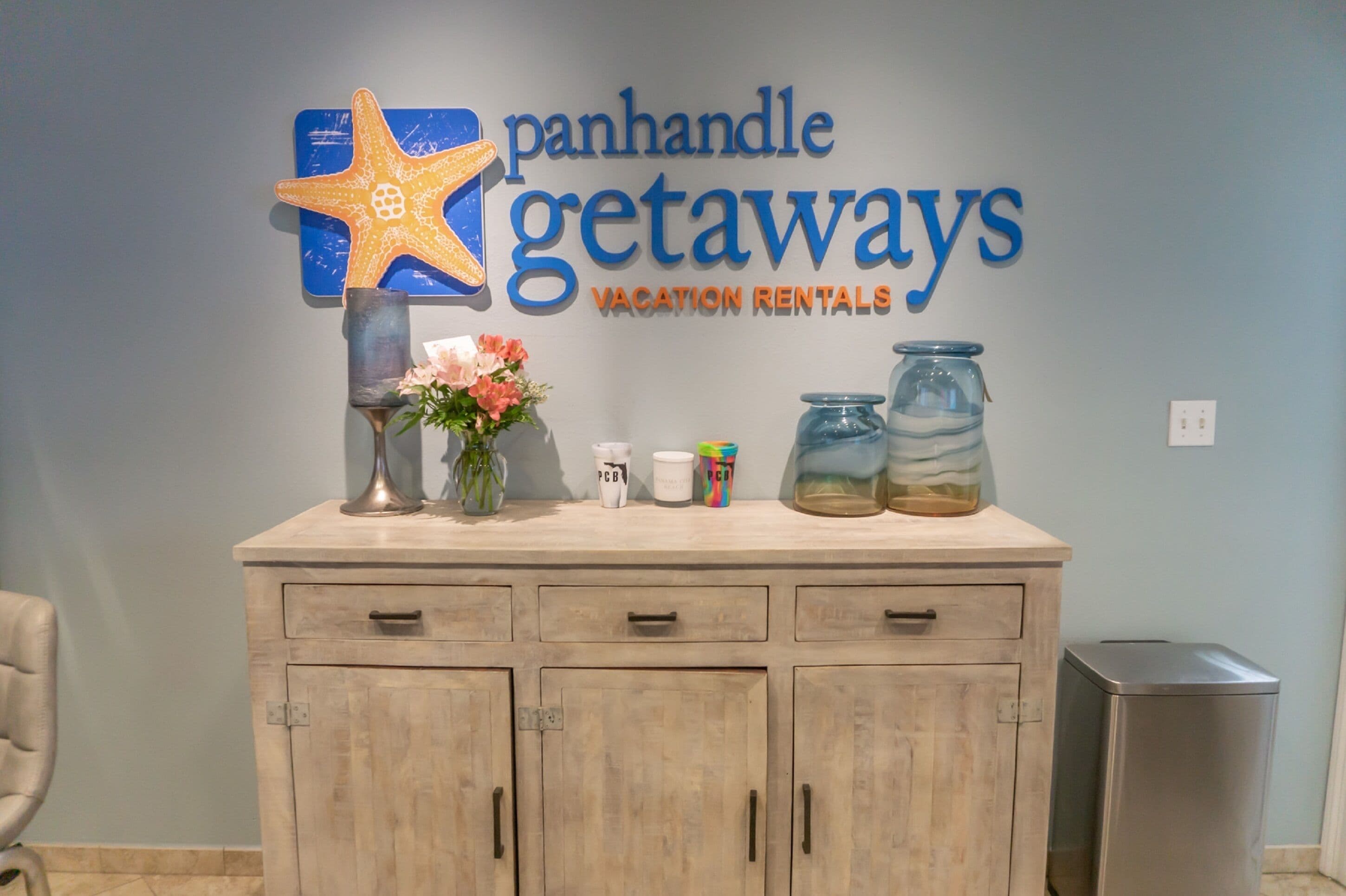 A reception desk with a sign reading "Panhandle Getaways Vacation Rentals," a starfish decoration, a vase with flowers, and two jars.