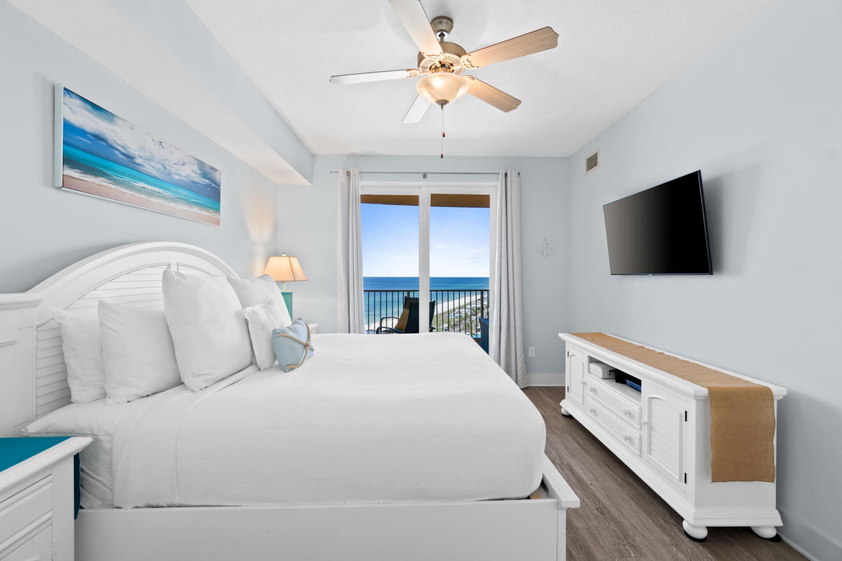 A bedroom with a bed, a ceiling fan, a TV, a window with a view of the ocean, and a white dresser.