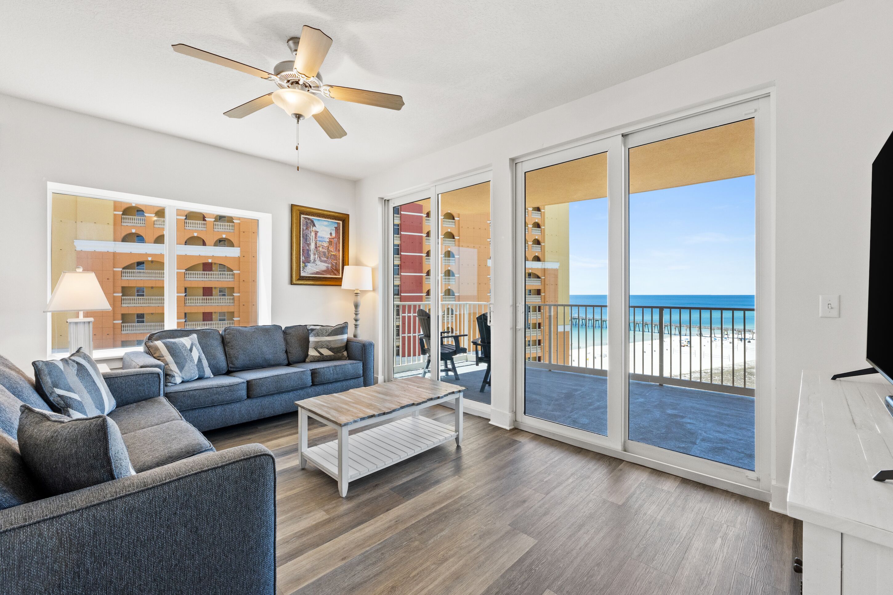 A spacious living room with a large sofa, a coffee table, and a ceiling fan. There are large windows offering a view of the beach and a balcony with a railing.