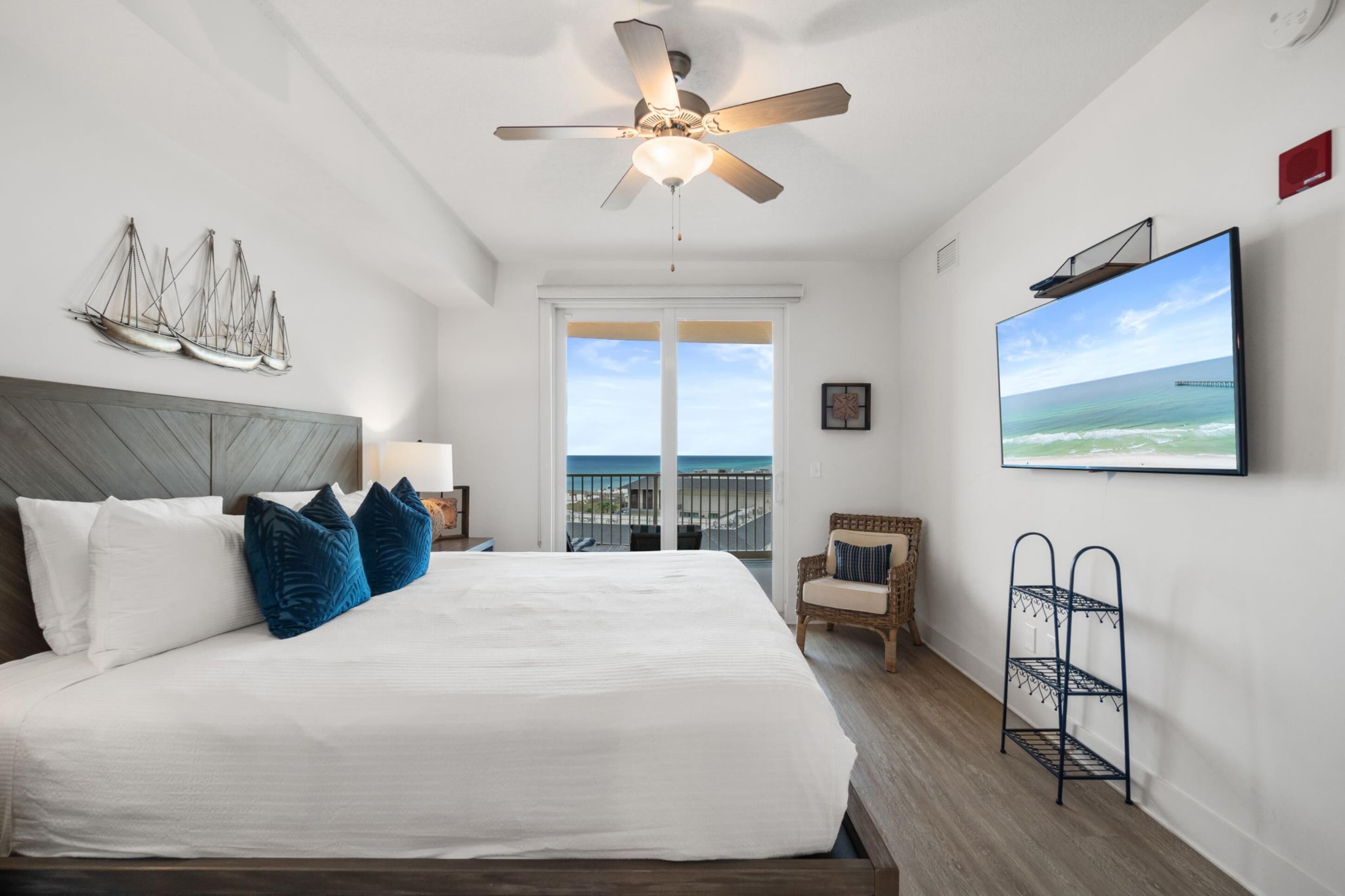 A hotel room with a large bed, a ceiling fan, a TV, a balcony with a view of the ocean, and a ship decoration on the wall.