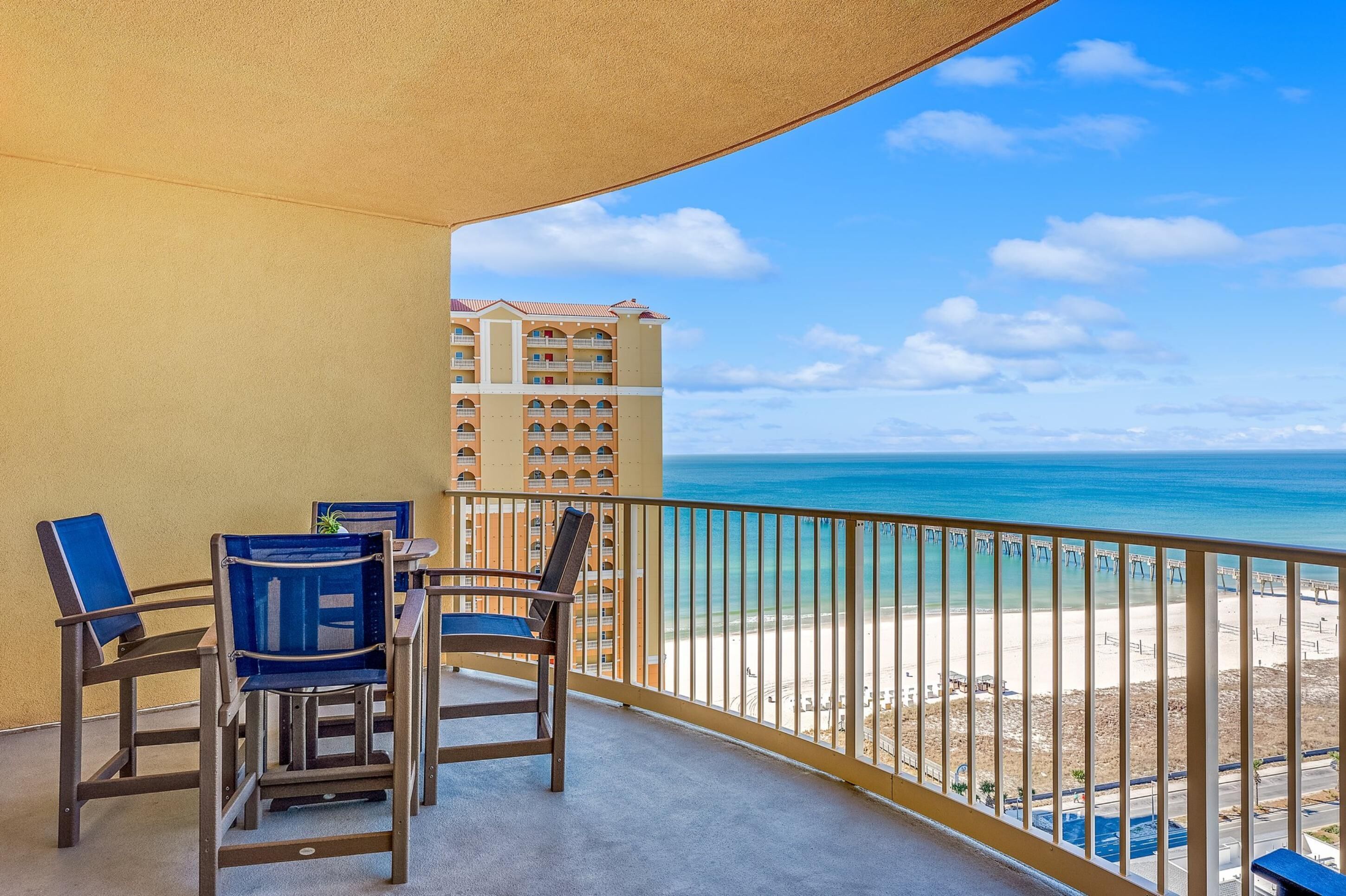 A balcony with two blue chairs and a table, overlooking a beach and a tall building.