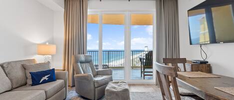 A hotel room with a sofa, armchair, ottoman, dining table, and a TV mounted on the wall.