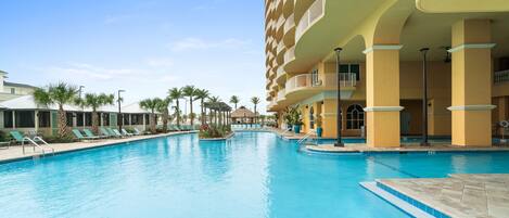 A hotel pool area with a clear blue pool, lounge chairs, and a multi-story building with balconies.