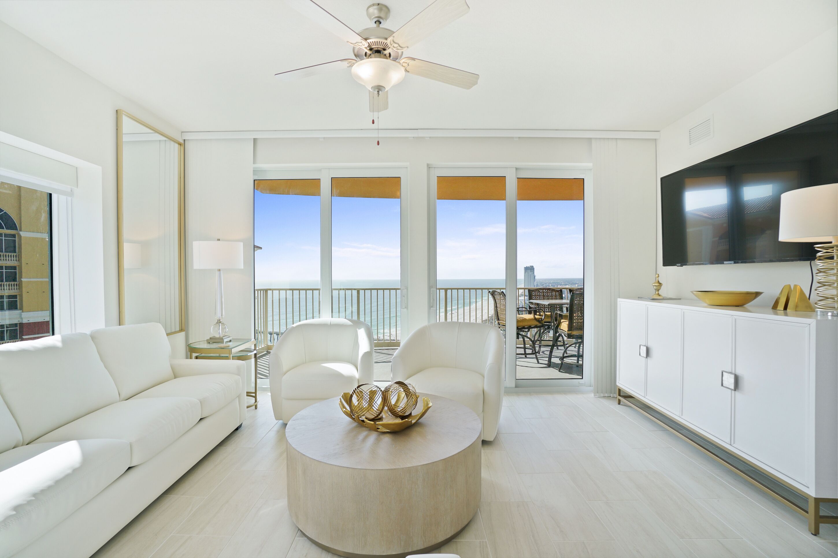 A spacious living room with a white sofa, two white armchairs, a round coffee table with decorative items, a ceiling fan, and a large TV on the wall.