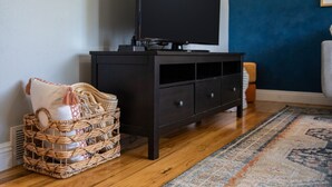 Sala de estar com um rack de madeira escura, uma cesta trançada com um travesseiro e um tapete estampado sobre piso de madeira.