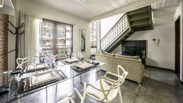 A dining area with a glass table, white chairs, and a sofa. A staircase leads to an upper level. - Loft Premium Imperdible (Ciudad Autónoma de Buenos Aires, Recoleta)