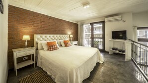 A bedroom with a brick wall, a white ceiling, a bed with white bedding, a nightstand, a lamp, a TV on a stand, and a balcony with a railing. - Loft Premium Imperdible (Ciudad Autónoma de Buenos Aires, Recoleta)