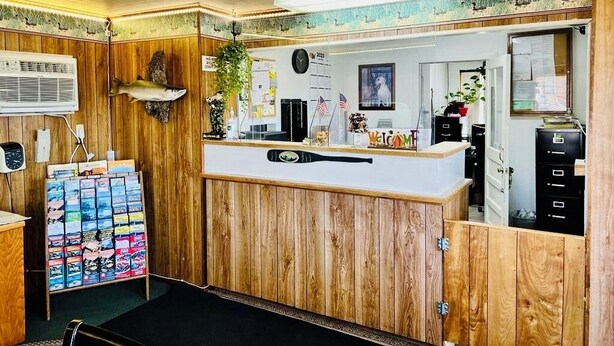 A reception area with a wooden counter, a clock, a plant, and a bulletin board with various items. - Sage N Sand Motel (Moses Lake)
