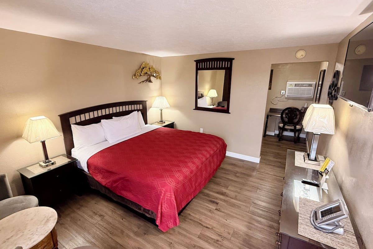 A hotel room with a bed, two bedside lamps, a desk with a telephone, a chair, a mirror, and an air conditioning unit.