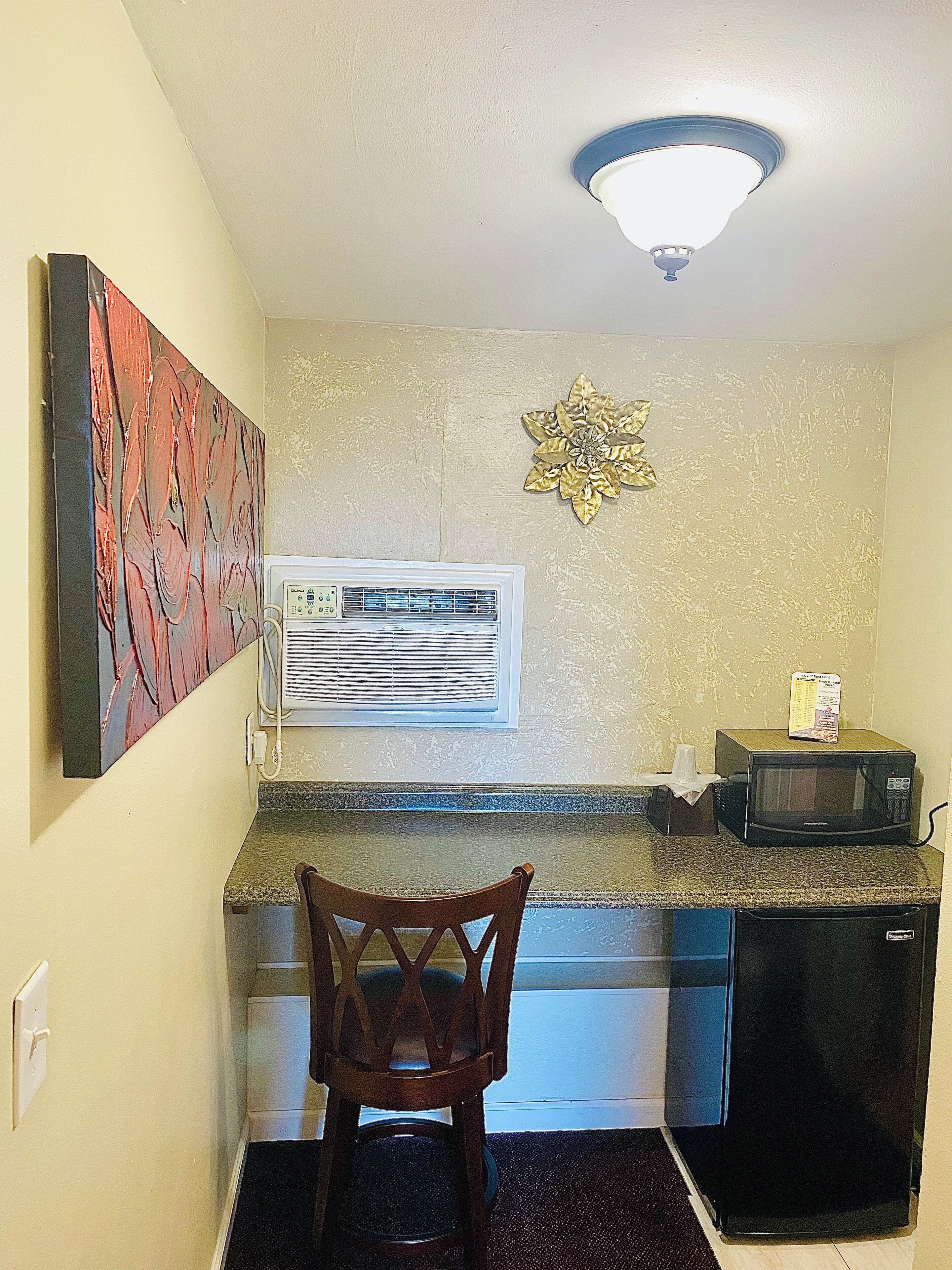 A compact kitchen with a microwave, a small refrigerator, a wall-mounted air conditioner, and a decorative wall piece.