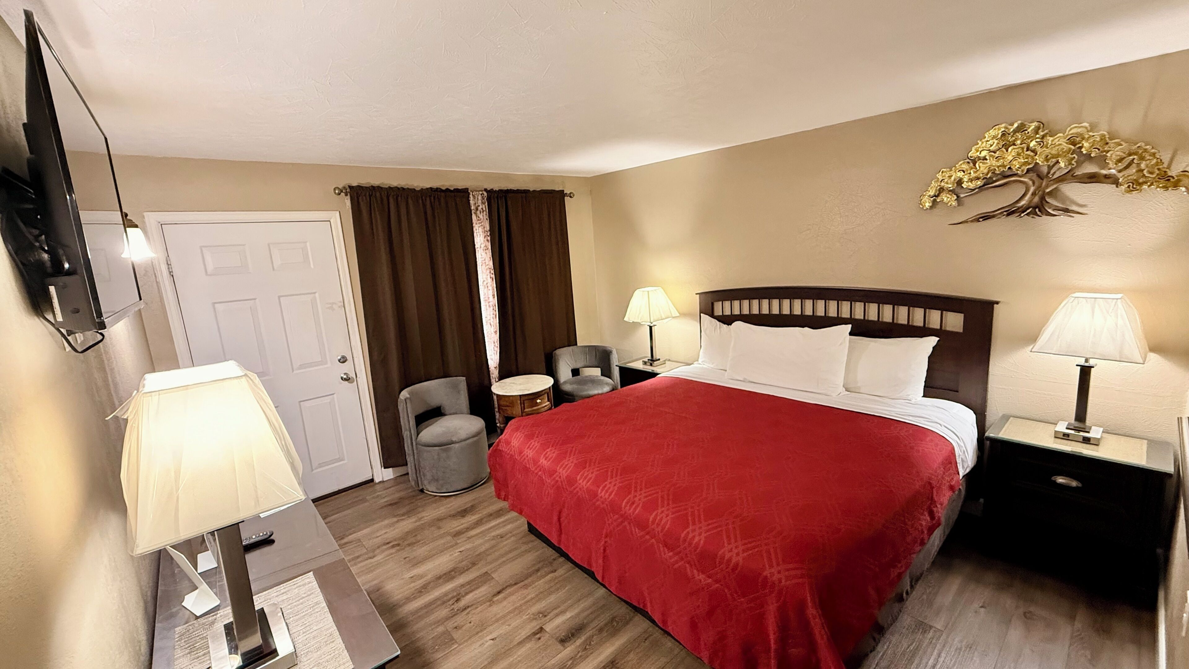 A hotel room with a bed, two bedside lamps, a chair, a small table, a TV, and a decorative tree on the wall.