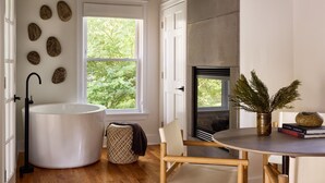 A modern bathroom with a large bathtub, a window with a view of greenery, a fireplace, and a round table with a vase and books. - AWOL Kennebunkport (Kennebunkport)
