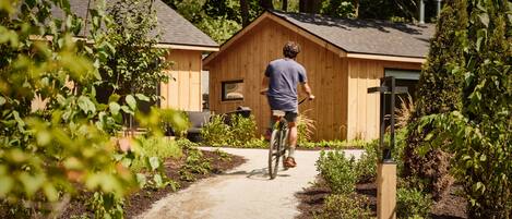 A person riding a bicycle on a dirt path towards a wooden cabin with a sloped roof, surrounded by greenery.