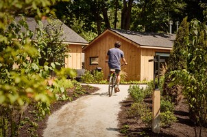 Una persona pedalea en bicicleta por un sendero de tierra hacia una cabaña de madera con techo inclinado, rodeada de vegetación.