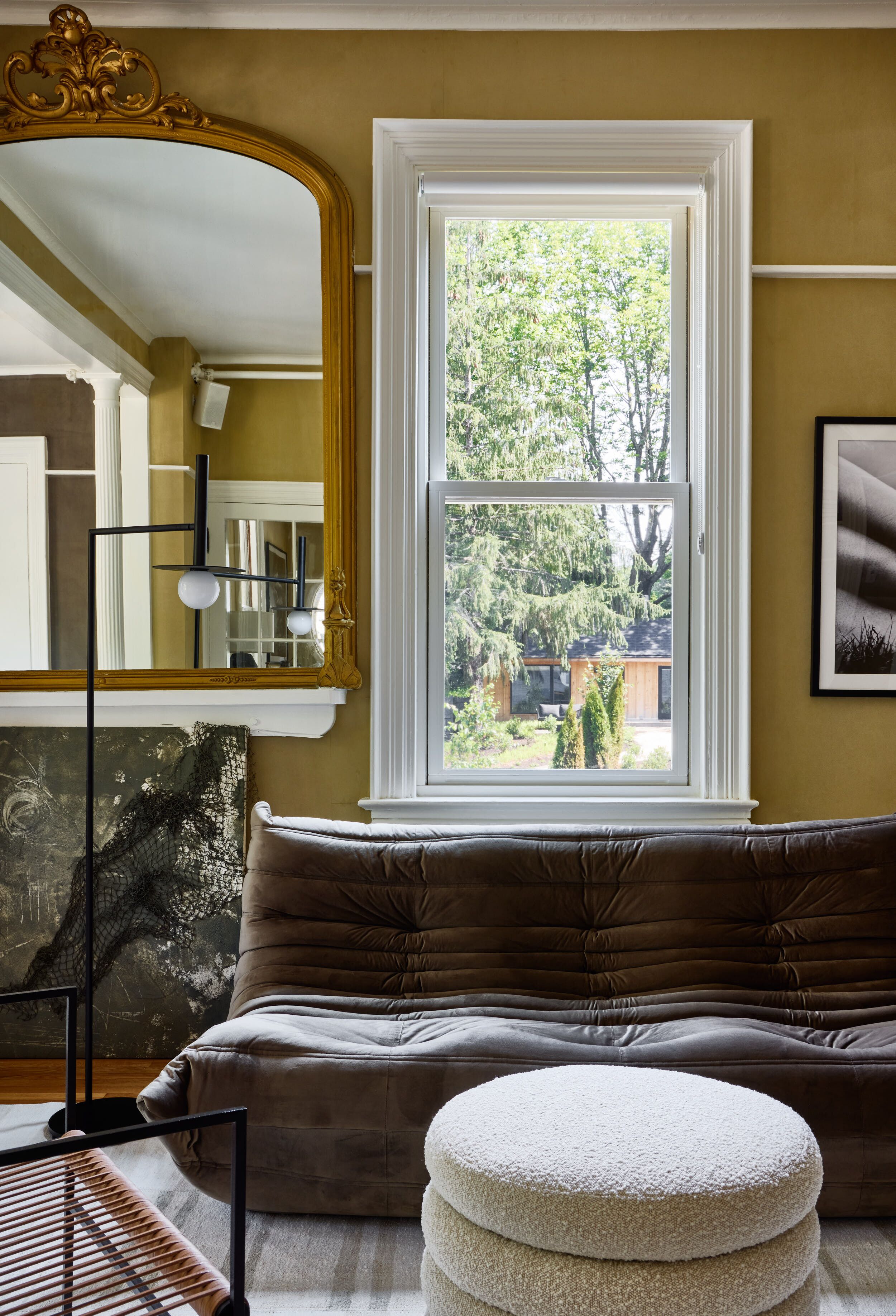 A room with a brown sofa, a round ottoman, a large mirror, and a window with a view of trees.