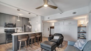 Una cocina moderna con una isla central, taburetes de bar, una butaca de cuero negro y un sofá gris.