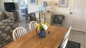 A dining area with a wooden table, white chairs, and a vase with flowers. There is a television in the background and a door with a view of a room with a bed and a lamp. - OCEANFRONT 2 BR CONDO! PANORAMIC OCEAN VIEWS FROM LIVING ROOM AND BEDROOMS  (Fort Pierce)