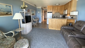 A living room with a sofa, a TV on a stand, a dining area with a table and chairs, and a kitchen with wooden cabinets and appliances.