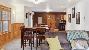 Ein Essbereich mit Holzsesseln und einem Tisch, ein braunes Sofa mit bunter Decke und eine Küche im Hintergrund.