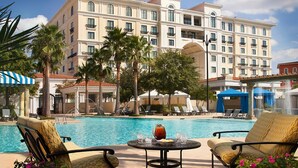 A hotel pool area with lounge chairs, a table with a drink, and a large building in the background. - EILAN HOTEL & SPA - 1 BR (San Antonio)