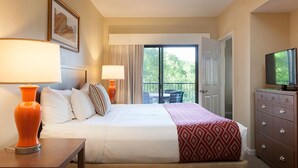 Habitación de hotel con cama, televisión, cómoda y balcón con vista a árboles.